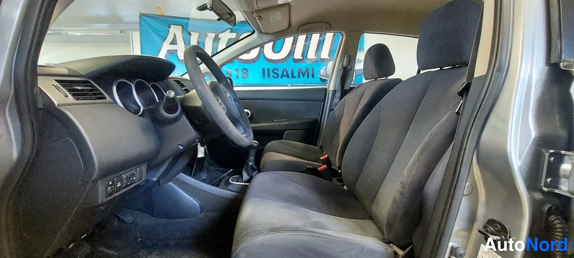 Muu: Nissan Tiidan ohjaamo sisältäpäin: tumma kangasverhoilu, manual-vaihde ja kojelauta.