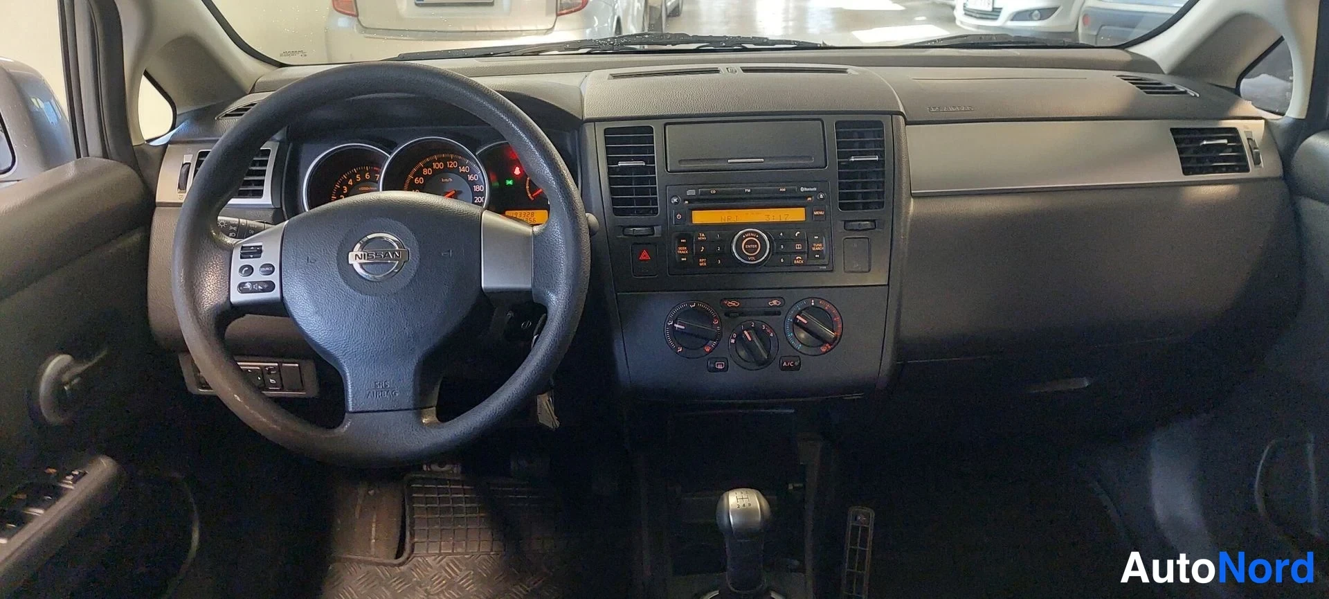 Muu: Ohjaamo Nikon Tiidasta: ohjauspyörä, mittaristo, radio ja manuaalivaihteen vaihdekeppi keskikonsolissa.
