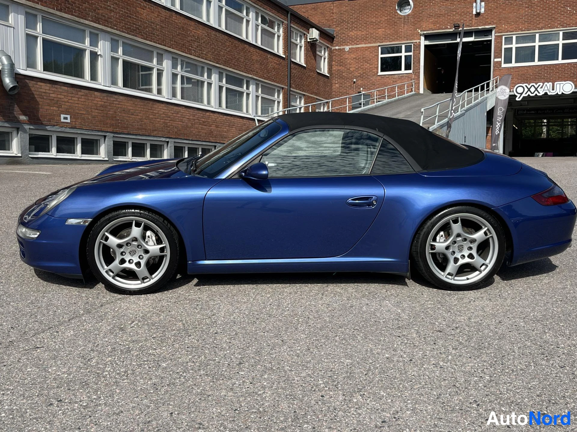 Etuviisto, nokka vasemmalle: Sininen Porsche 911 Cabriolet sivukulmassa nenä vasemmalla, musta katto, hopeiset kevytmetallivanteet.