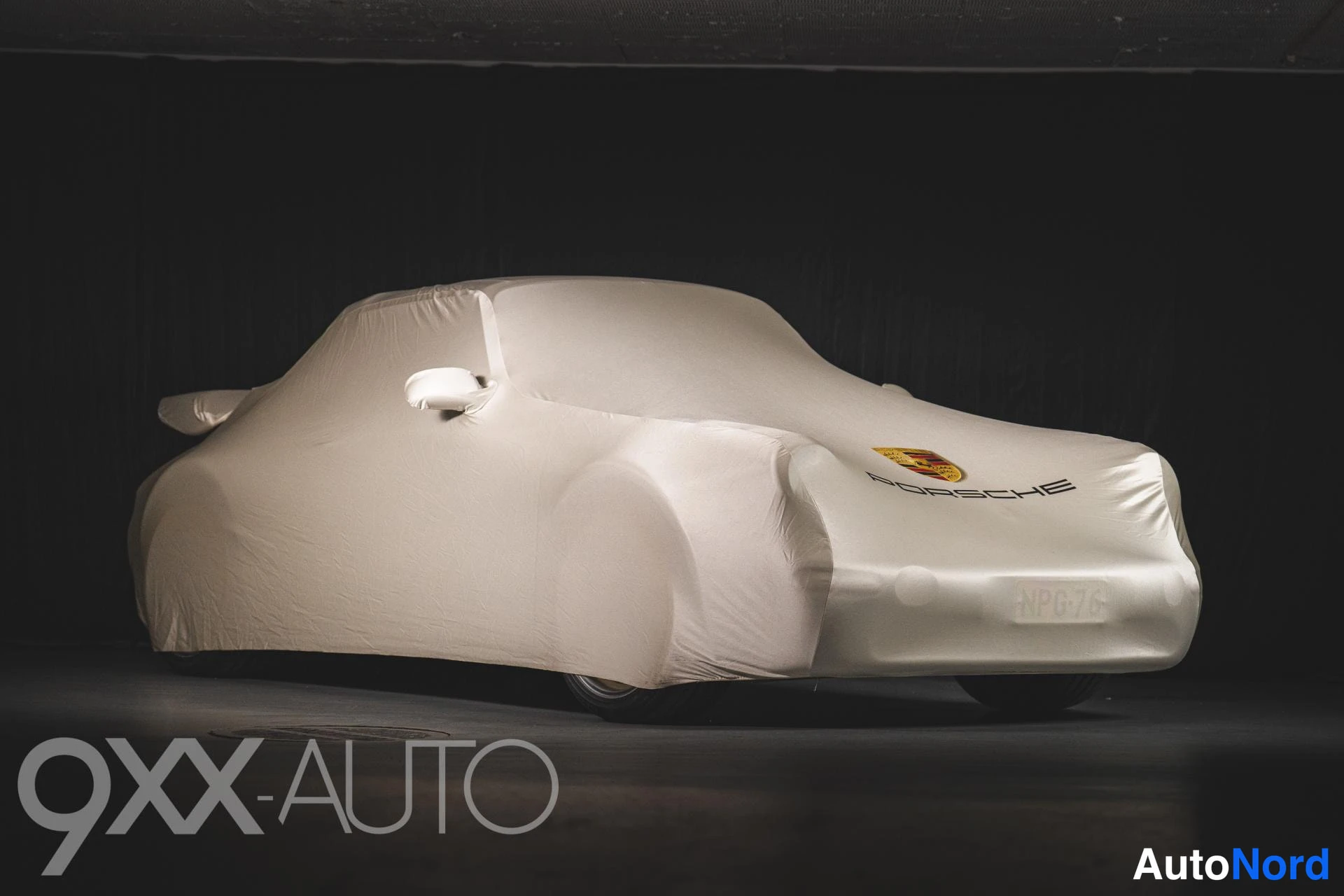 Etuviisto, nokka oikealle: Auto suojapeitteessä studiossa: peitteen alla erottuu takasiipi ja keula, jossa Porsche-logo.
