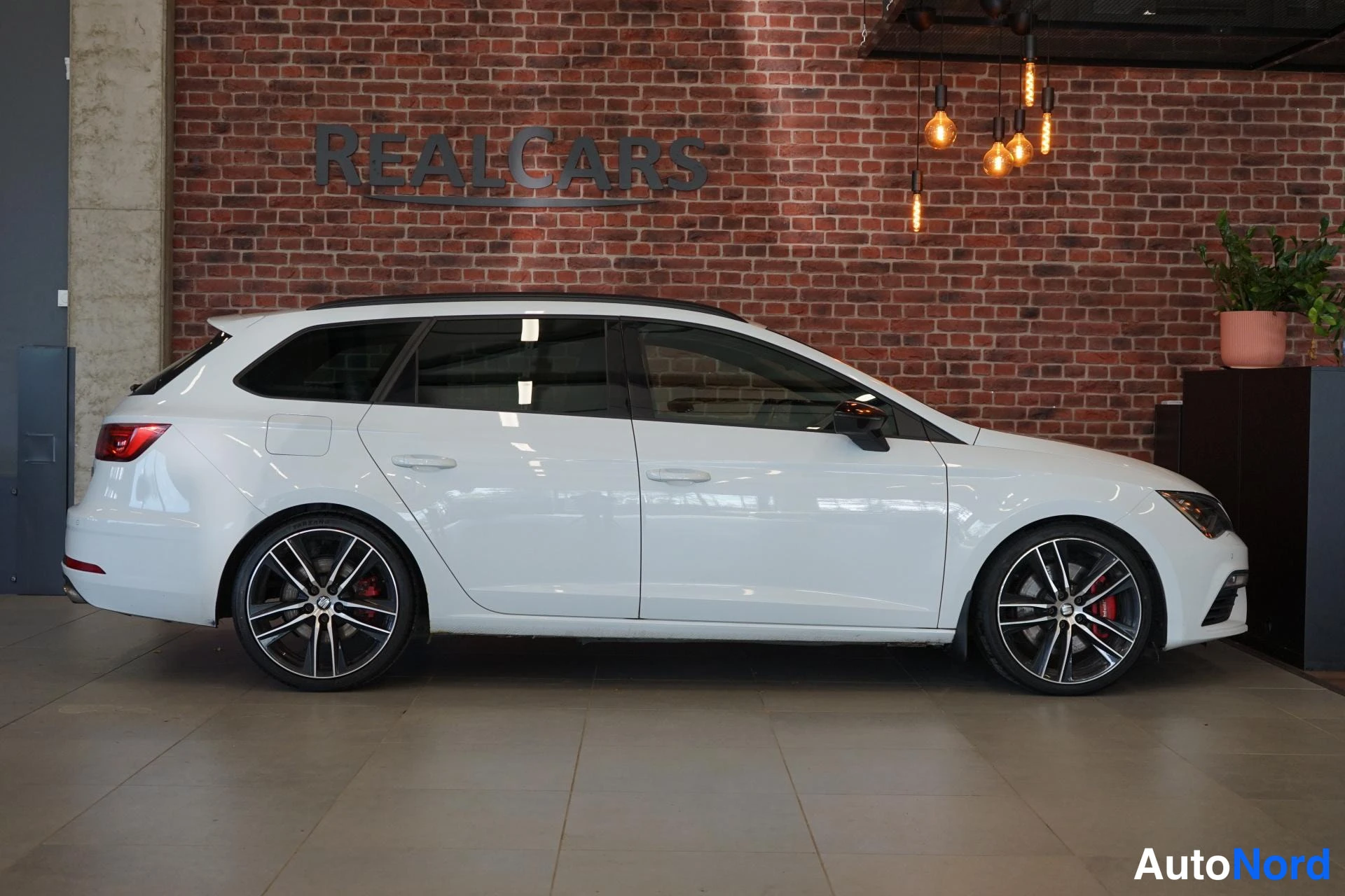 Sivulta: Valkoinen Seat Leon ST -farmari sivusuunnasta sisätiloissa tiiliseinää vasten.
