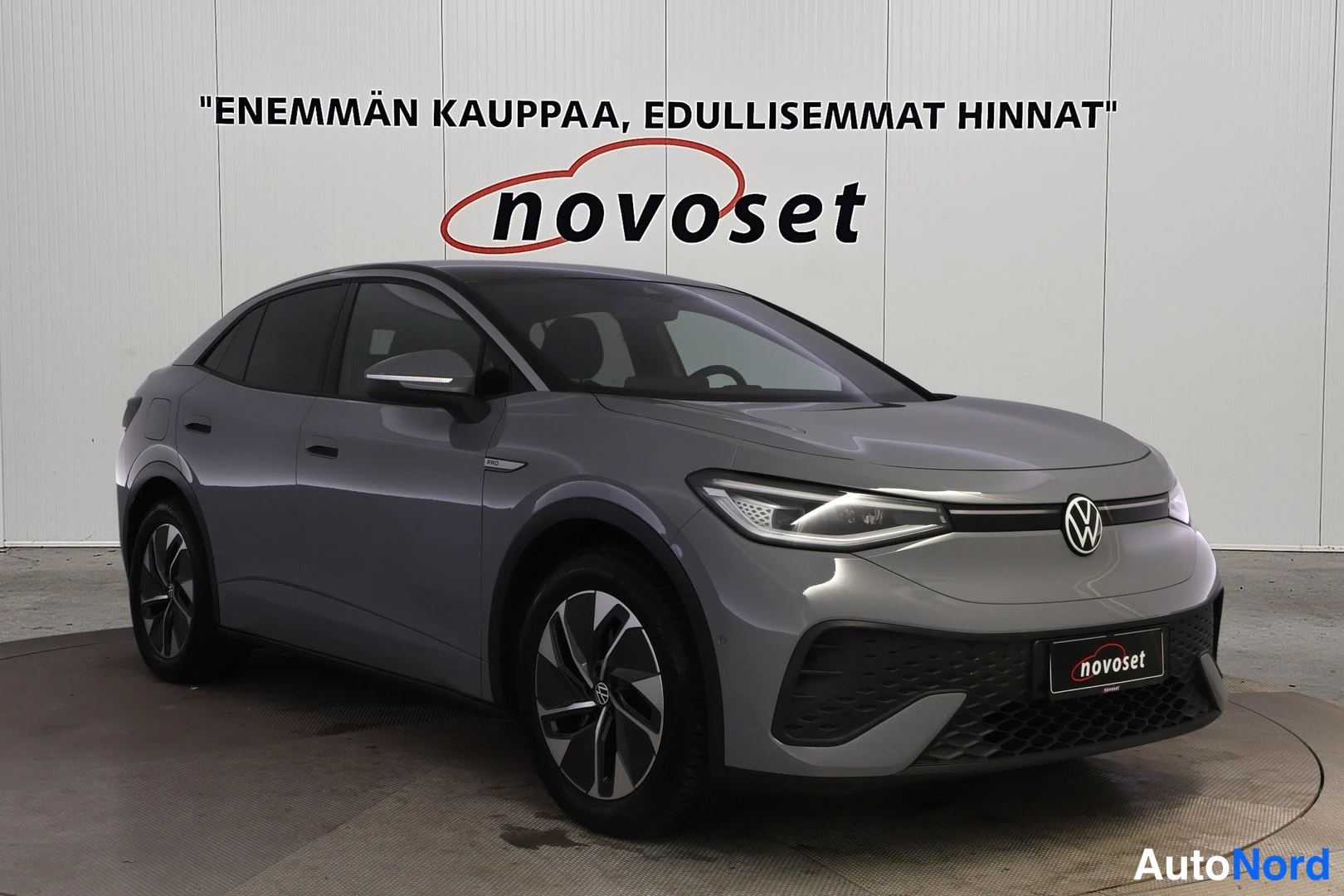 Etuviisto, nokka oikealle: Auton etukulmakuva: harmaa Volkswagen-henkinen sähköauto kylttiä novoset vasten, keulakilpi ja vasen sivu näkyvissä.