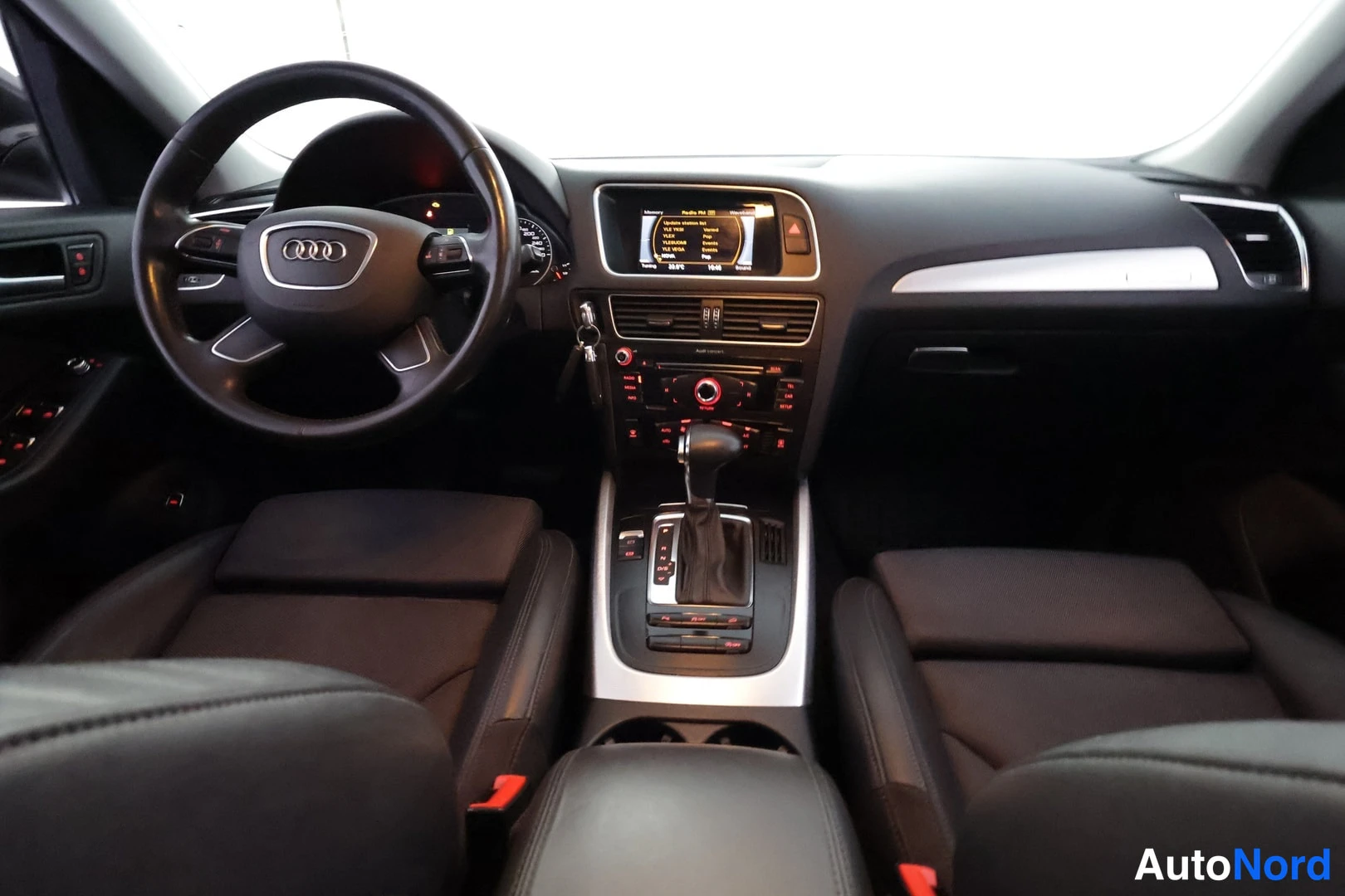 Muu: Ohjaamo kuvattuna etupenkkien yli: Audi-ohjauspyörä, kojetaulin näyttö, keskikonsolin säätimet ja automaattivaihteen vipu.