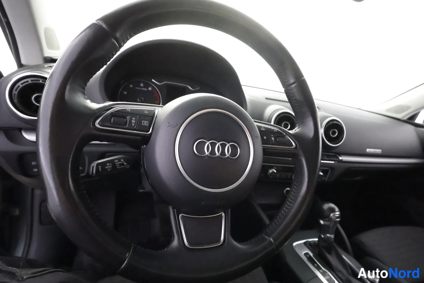 Muu: Sisäkuva: musta ohjauspyörä, jonka keskussa on Audi-logotunnus, kojelauta ja automaattivaihteen valitsin näkyvät taustalla.