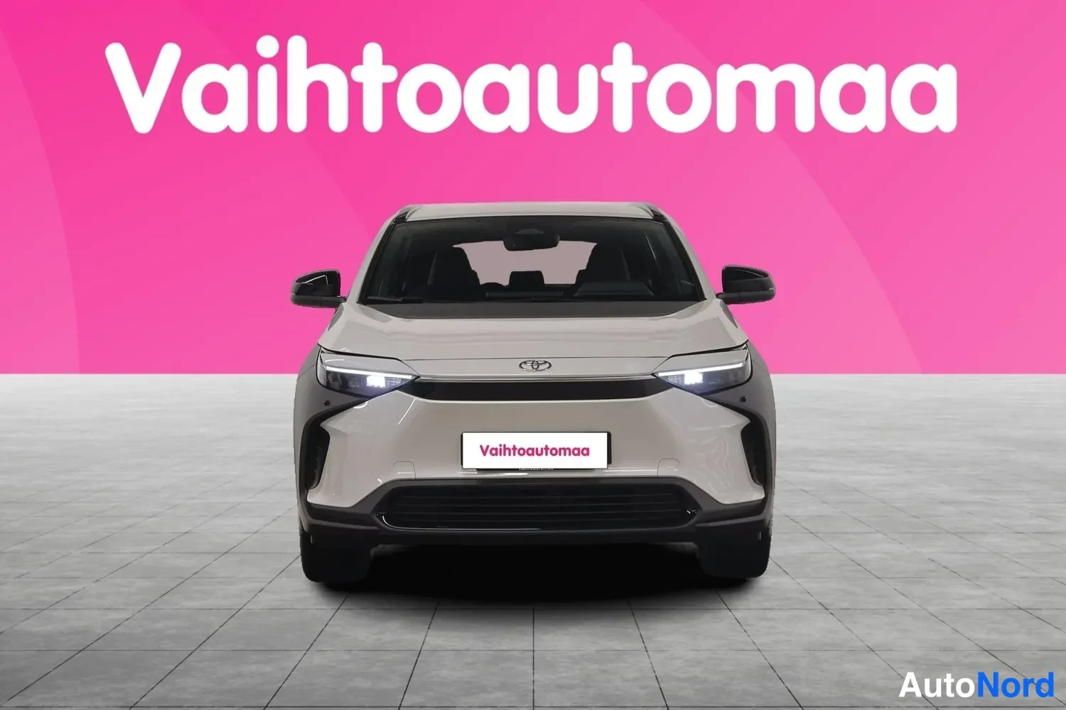 Edestä: Toyota bZ4X -sähköauto suoraan edestä kuvattuna, LED-ajovalot päällä, etupuskurissa Vaihtoautomaan tunnuskilpi.