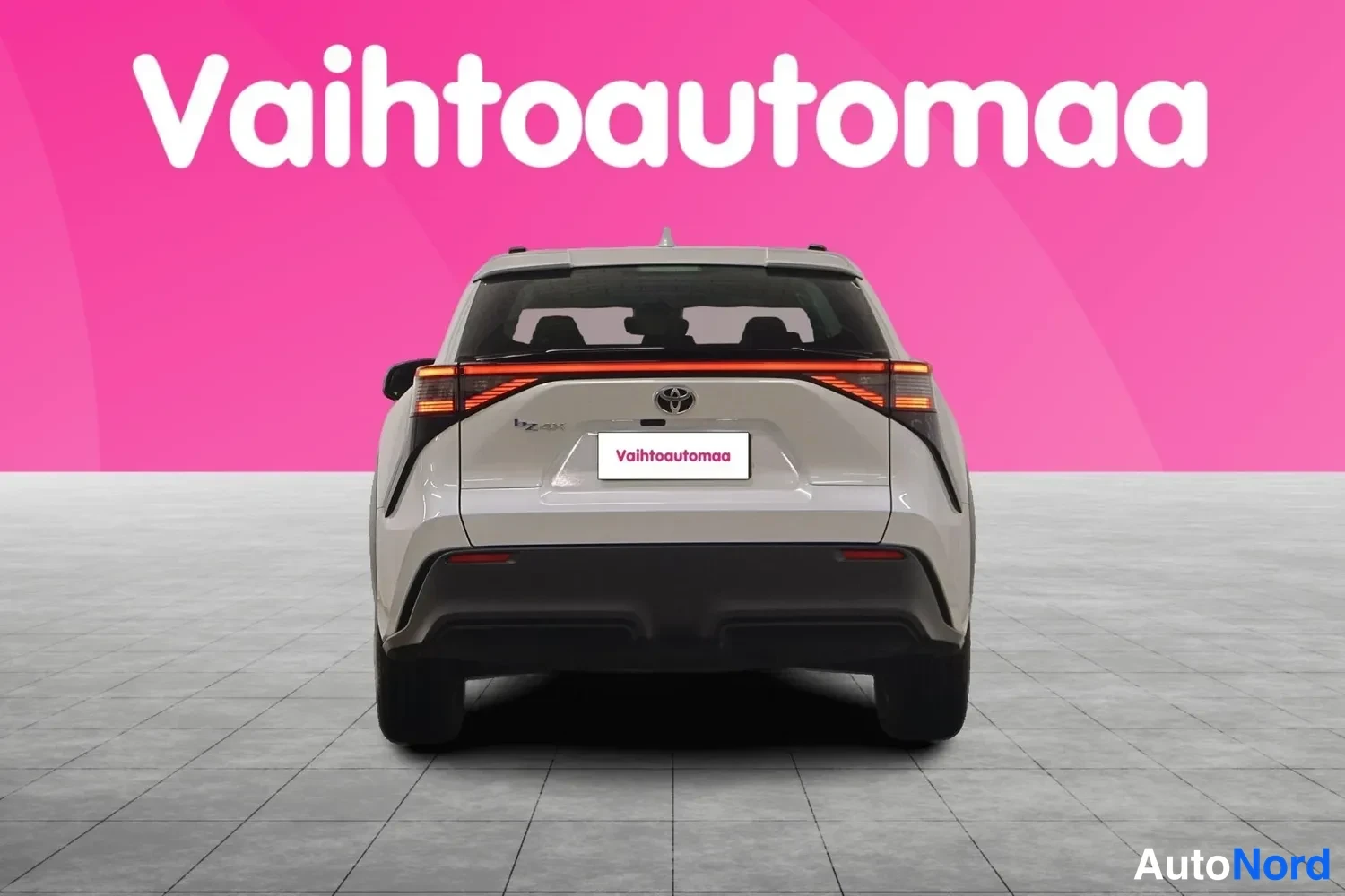 Takaa: Auton takaosa suoraan takaa: Toyota-merkki ja vaalea koriväri, rekisterikilven paikalla mainoslaatta.
