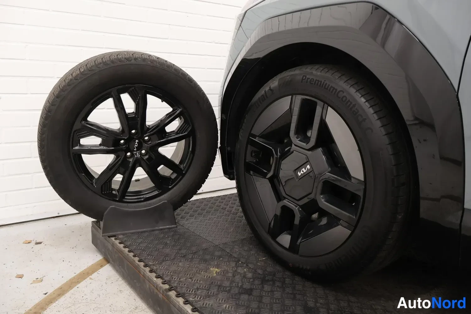 Muu: Kia-merkitty musta kevytmetallivanne ja sen vieressä irrotettu varapyörä mustalla vanteella, Continental PremiumContact 5 -rengas.