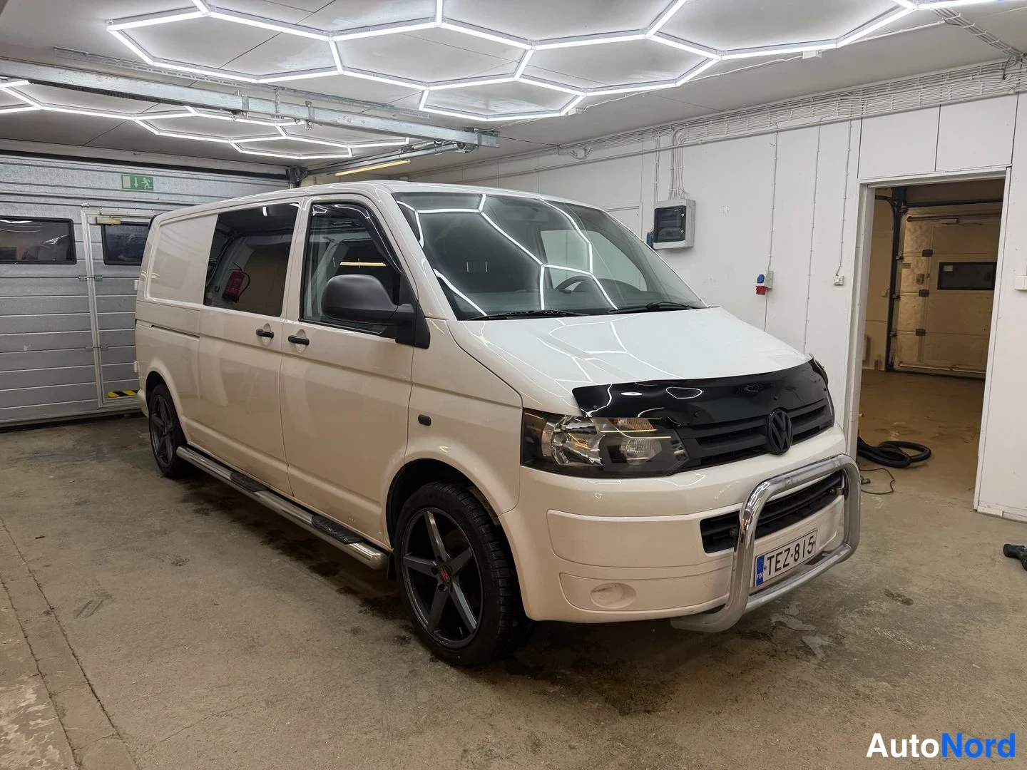 Etuviisto, nokka oikealle: Valkoinen Volkswagen Transporter -pakettiauto etu-nurkkakuva sisätiloissa, katse oikeaan; etusuoja, keulakaari ja tummat vanteet näkyvät.