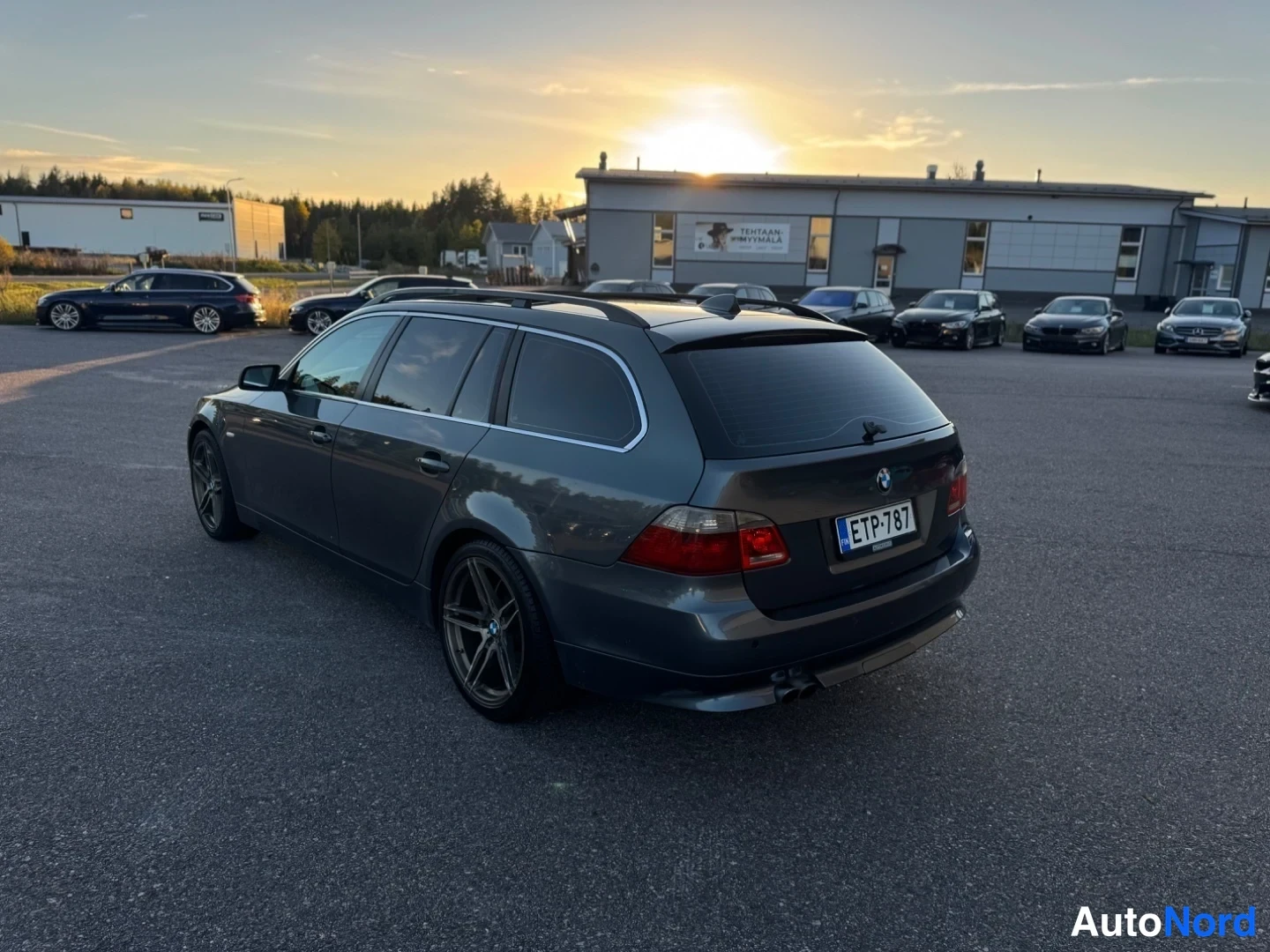 Takaviisto: Tummanharmaa BMW 5-sarjan farmari kulmatrista takaa vasemmalta auringonlaskun valossa, rekisteritunnus.