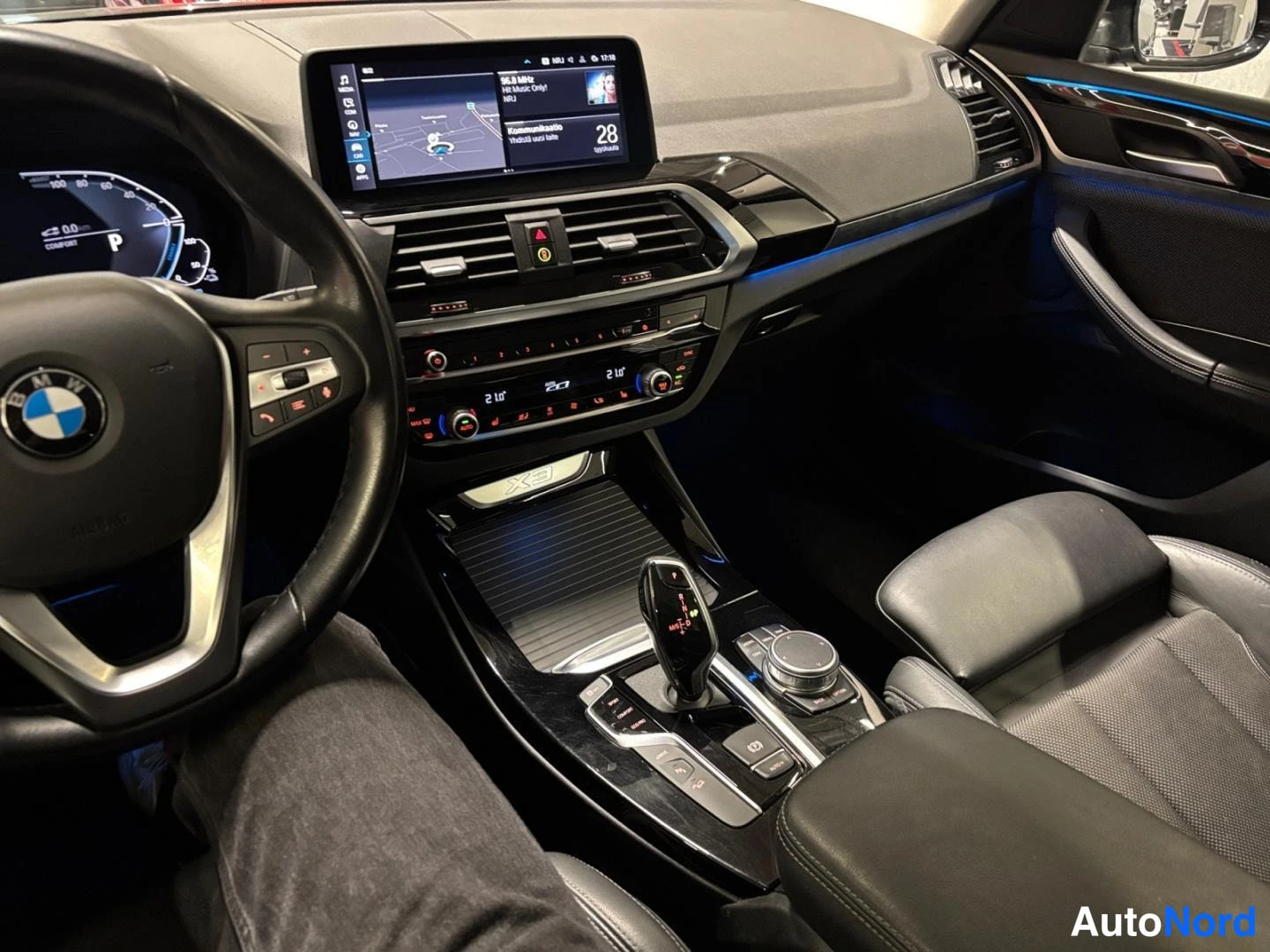 Muu: Ohjaamo näkymä BMW-merkillä ratissa, keskellä kosketusnäyttö karttanäkymällä, automaattivaihteiston valitsin ja keskikonsolin säätimet.