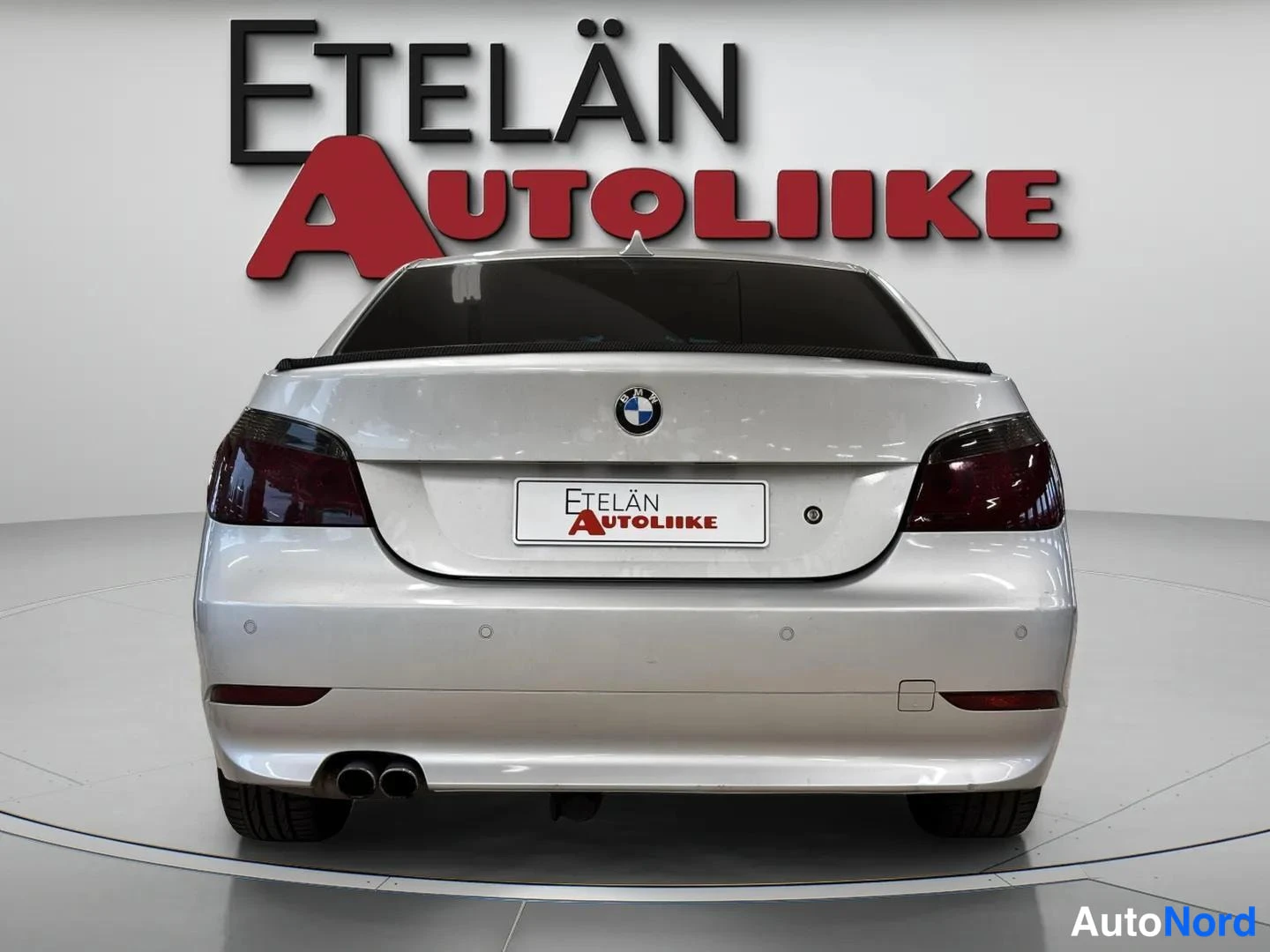 Takaa: Auton suora takanäkymä: hopeanvalkoinen sedan, BMW-merkki kannessa, tummat takavalot, Etelän Autoliike-kyltti rekisterin paikalla ja kaksoispakoputki vasemmalla