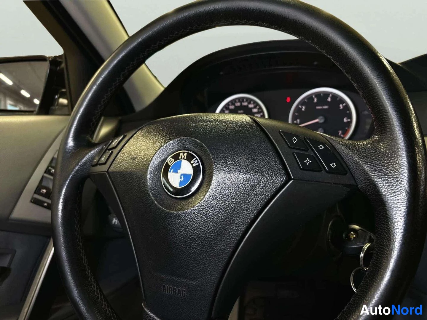 Muu: BMW-ohjauspyörä lähikuvassa: keskussa BMW-embleema, AIRBAG-teksti alaosaan ja monitoimapainikkeet oikealla puolella; takana osittain näkyvät mittarit.