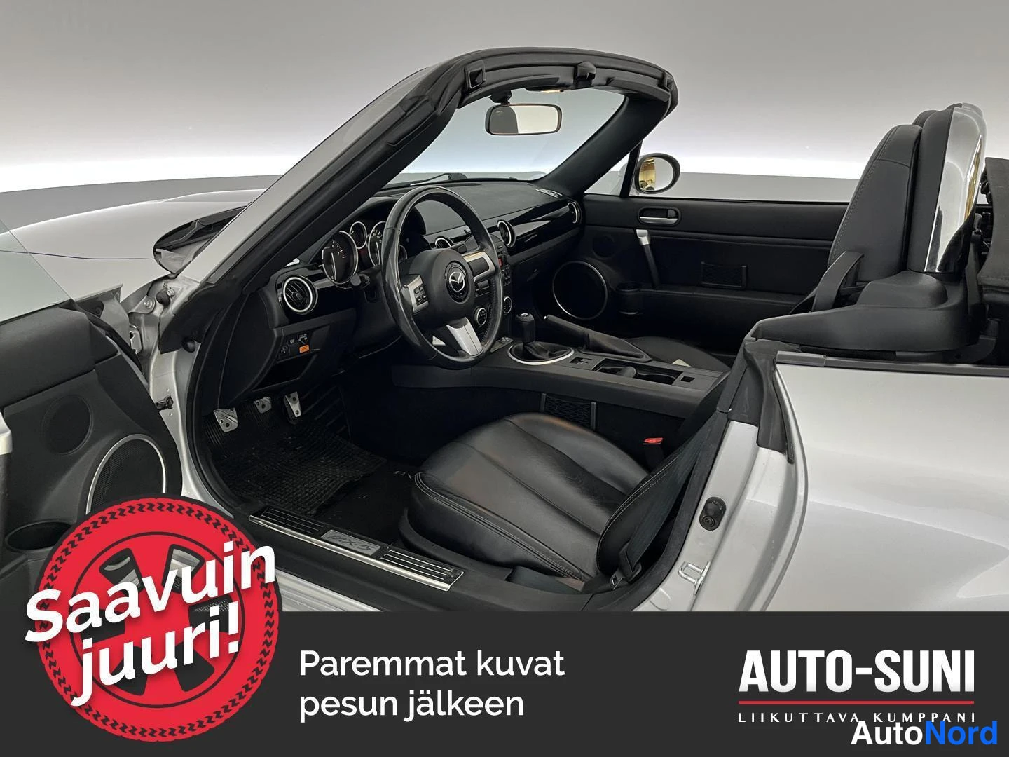 Etuviisto, nokka oikealle: Avauskuva Mazda MX-5:n ohjaamosta: musta nahkainen sisusta, ohjauspyörä, kojelaudan mittarit ja avattu pehmeä katto.
