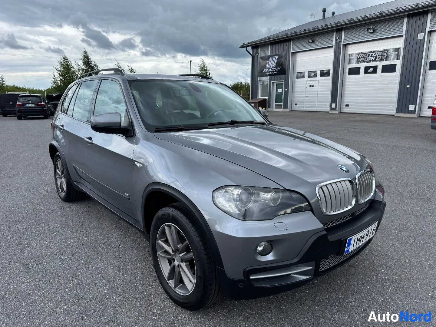Etuviisto, nokka oikealle: Hopeanharmaa BMW X5 etuviistosta oikealta, rekisterikilpi 1MM-916, parkkihalli autotallirakennuksen edessä.