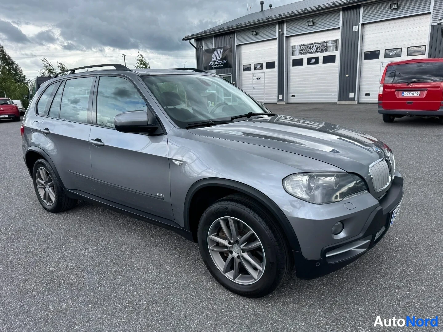 Etuviisto, nokka oikealle: Hopeanharmaa BMW X5 etuviistosta oikealta sivukulmassa, katuympäristö ja punainen pakettiauto taustalla.