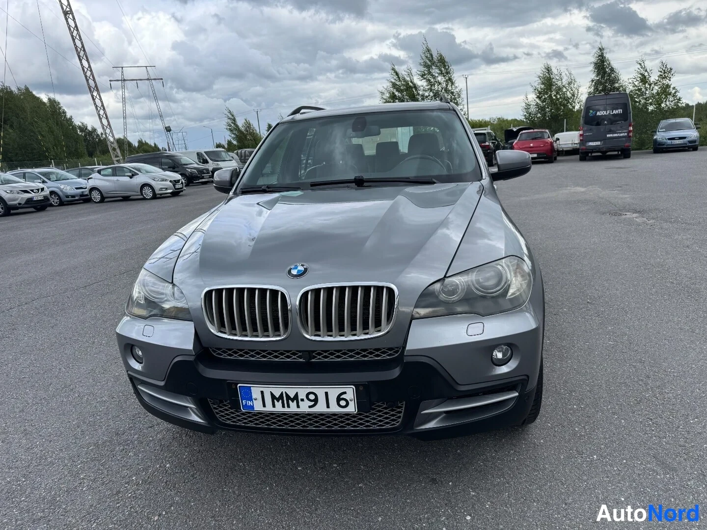 Edestä: Hopeanharmaan BMW X5:n etupää suoraan edestä, rekisterikilpi 1MM-916, parkkialue muiden autojen kanssa.