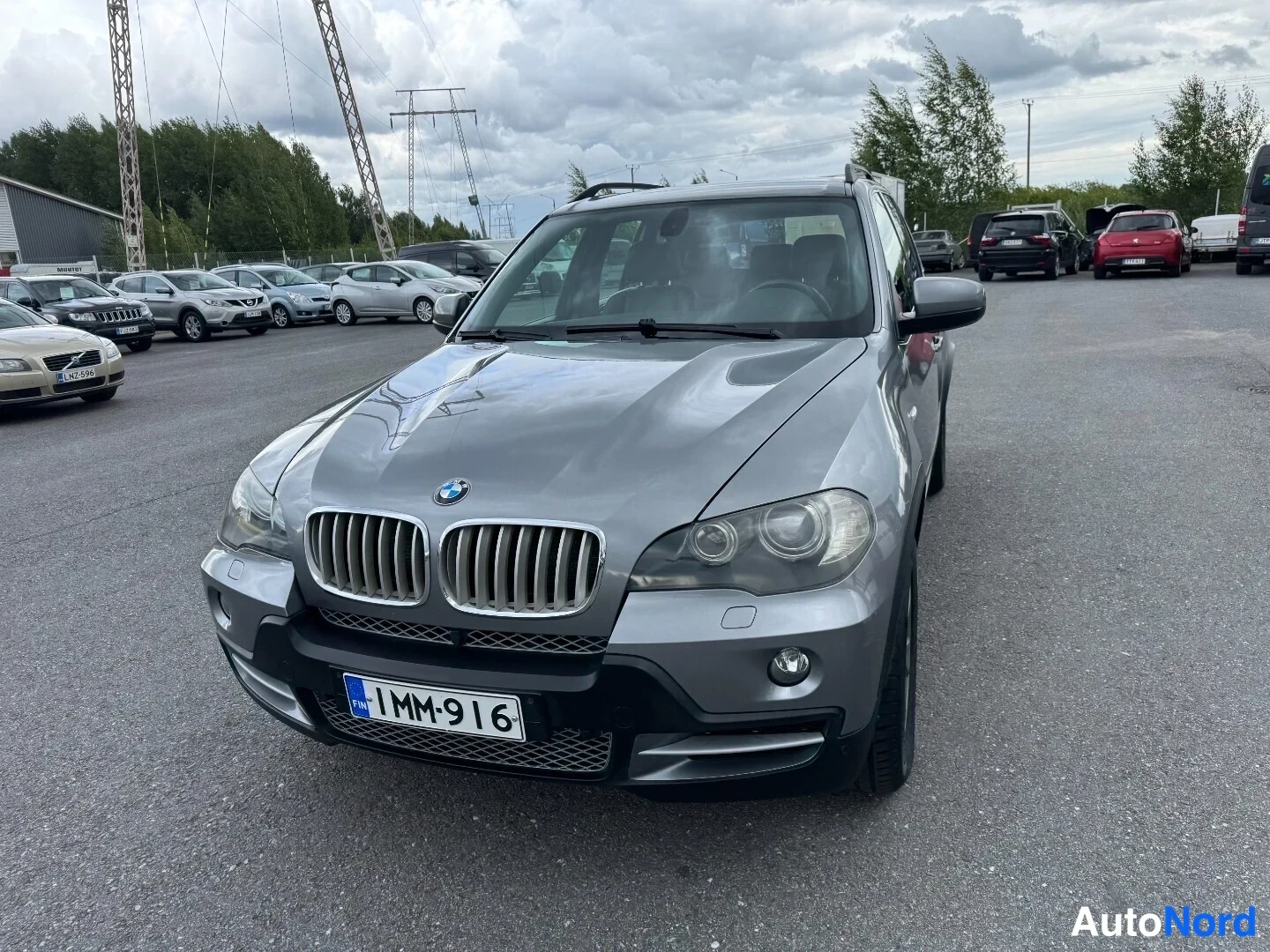 Edestä: BMW X5:n etupää hieman vinosti edestä, rekisterikilpi 1MM-916, parkkialue pilvisellä säällä.