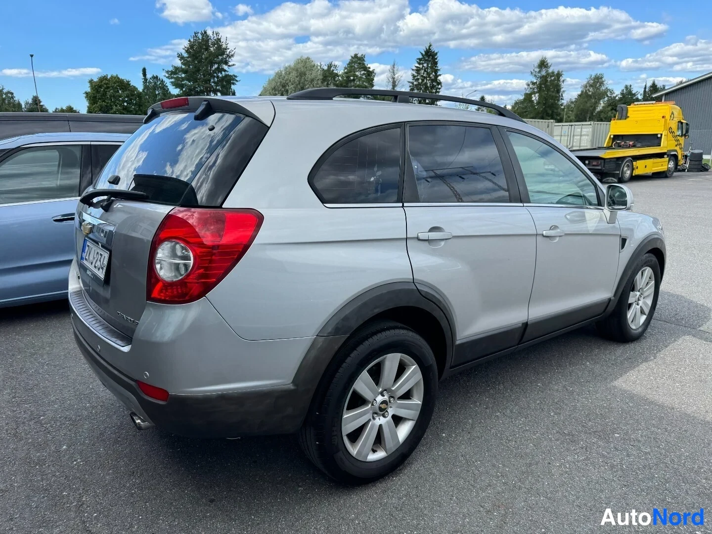 Takaviisto: Chevrolet Captiva takaosan ja vasemman sivun näkyvissä; hopeinen koripinta, kattokaiteet ja viisivaiheiset pyöränkannattajat näkyvät.