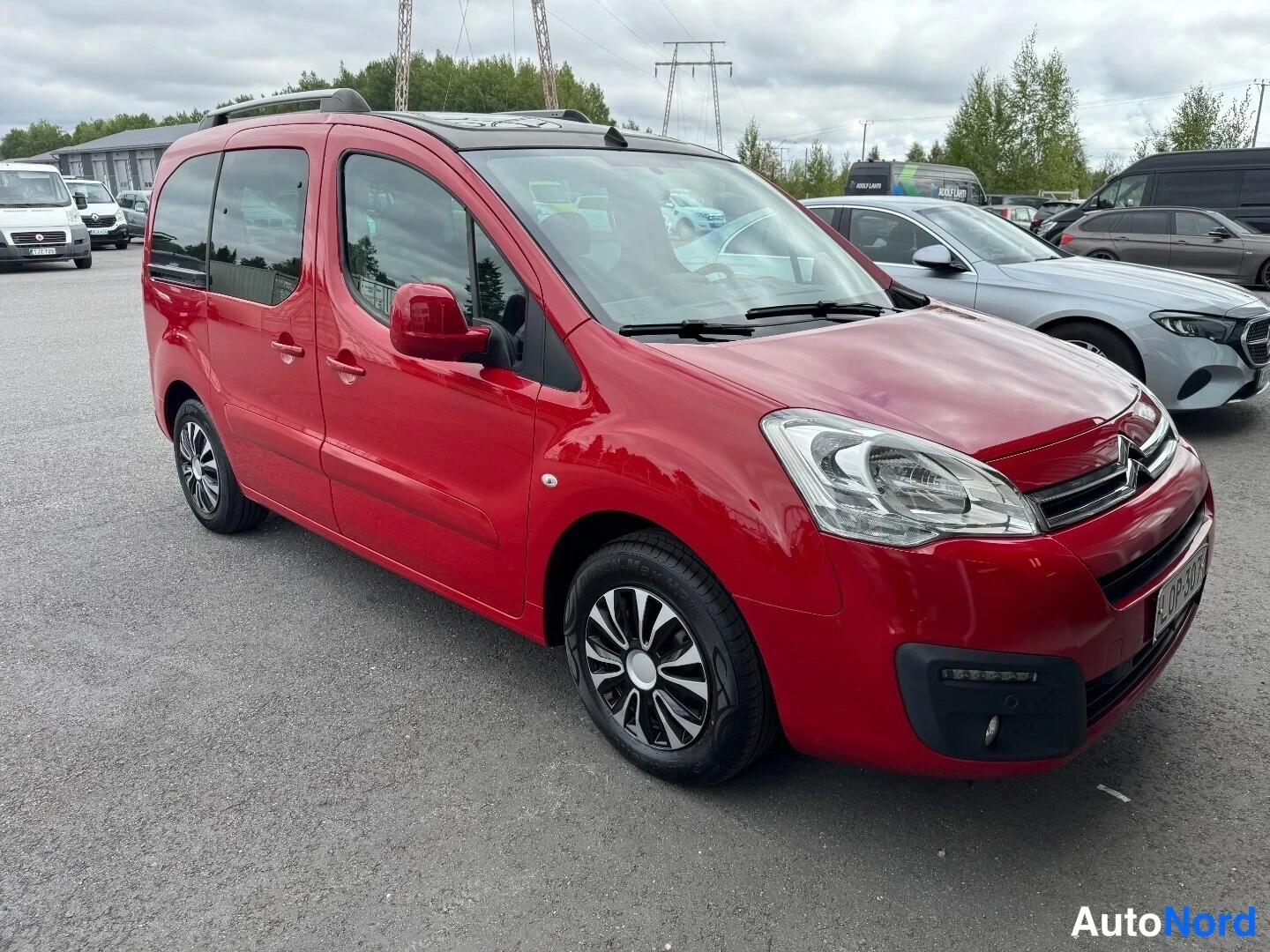 Etuviisto, nokka oikealle: Punainen Citroen Berlingo -pakettiauto vasemman kyljen ja keulan näkymässä parkkipaikalla.