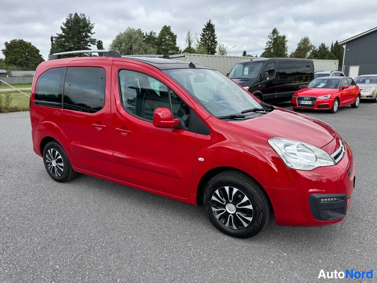 Etuviisto, nokka oikealle: Punainen Citroen Berlingo -pakettiauto sivulta edestä kuvattuna, tumma lasikatto ja kattokaiteet.