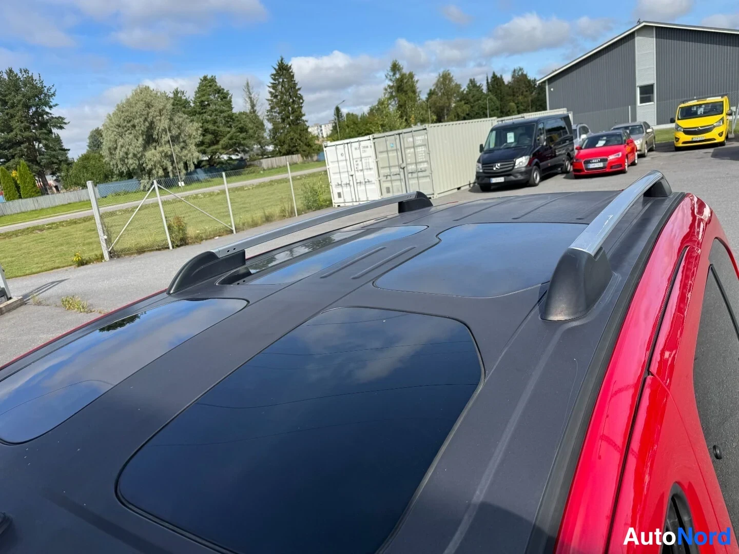 Muu: Kattotäydellinen harmaa lasipaneeli ja kattokaiteet punaisessa korissa; taustalla pihamaa, kontteja ja muita ajoneuvoja.