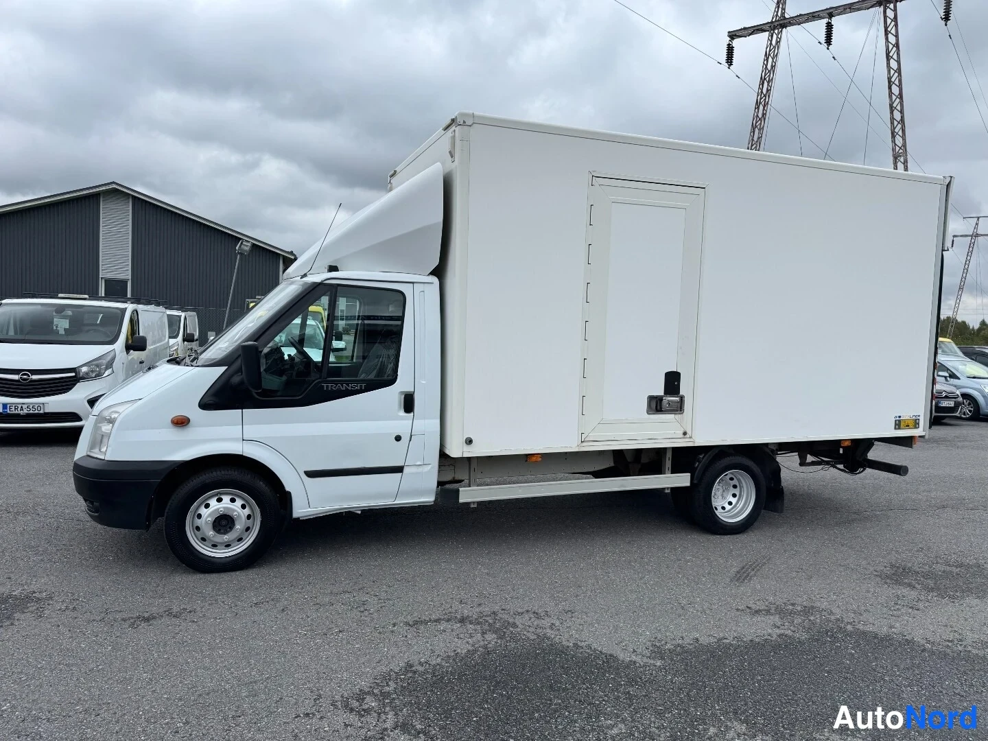 Etuviisto, nokka vasemmalle: Valkoinen Ford Transit kuorma-auto nokka vasemmalla, sivunosto-ovi takaosassa.
