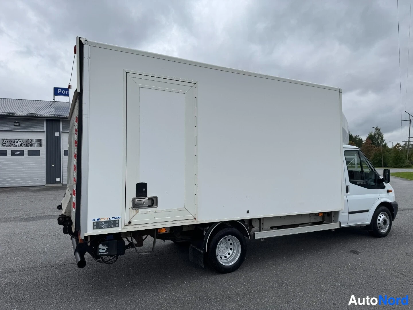 Takaviisto: Valkoinen Ford Transit takaosa vasemmalla, Perkins-lavajärjestelmä näkyvissä takaoven kohdalla.