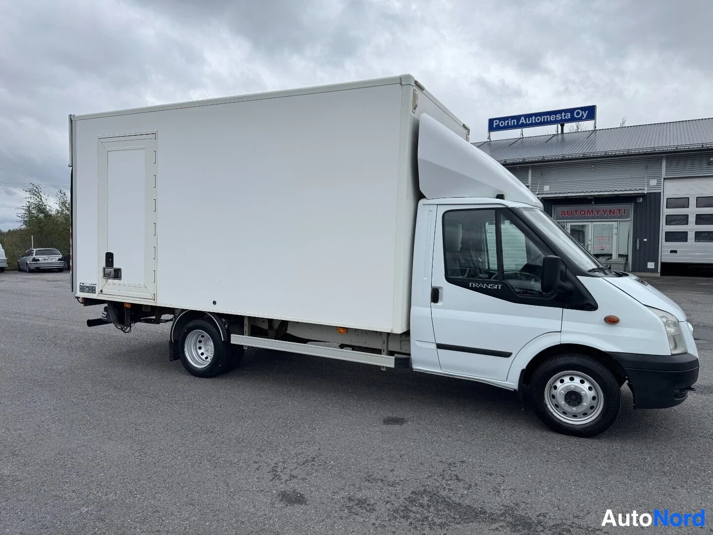 Etuviisto, nokka oikealle: Valkoinen Ford Transit-kuorma-auto sivulta etuviistoon, TRANSIT-teksti ovessa ja Porin Automesta Oy -kyltti taustalla.