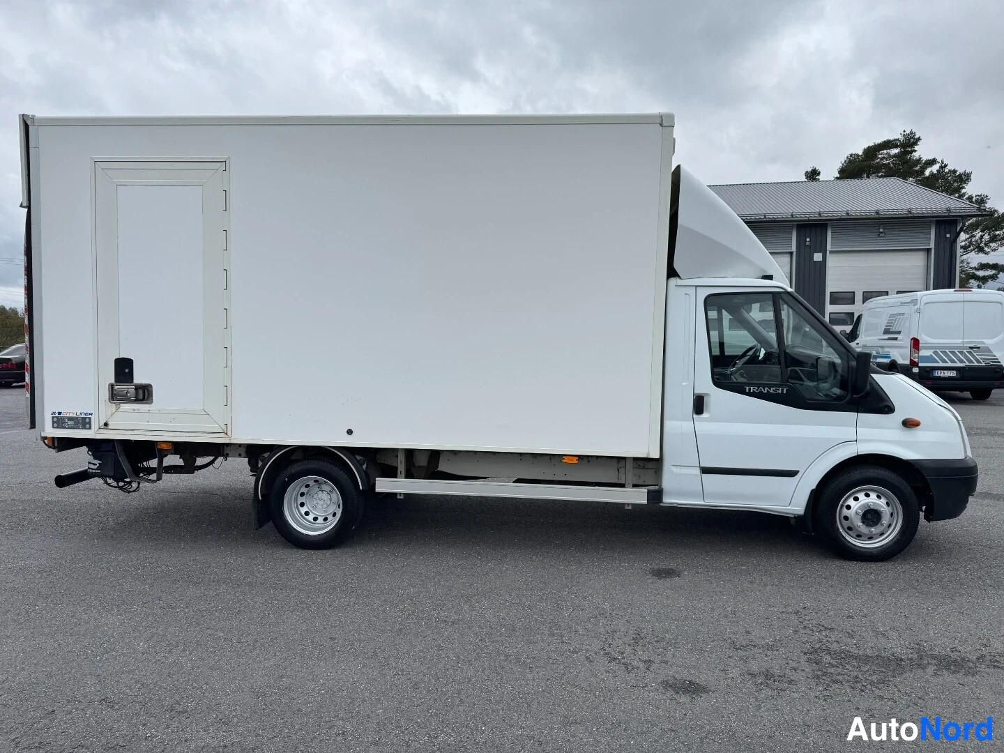 Sivulta: Valkoinen Ford Transit-kuorma-auto kokonaan sivusta, TRANSIT-teksti ovessa ja valkoinen lavaosasto.