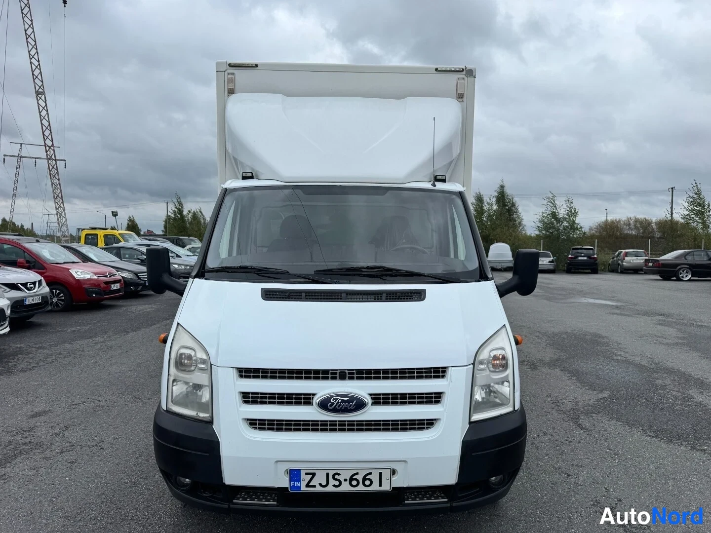 Edestä: Valkoisen Ford Transit -kuormaauton etunäkymä, valkoinen lavettipohjainen pakettiauton korirakenne, rekisterikilpi.