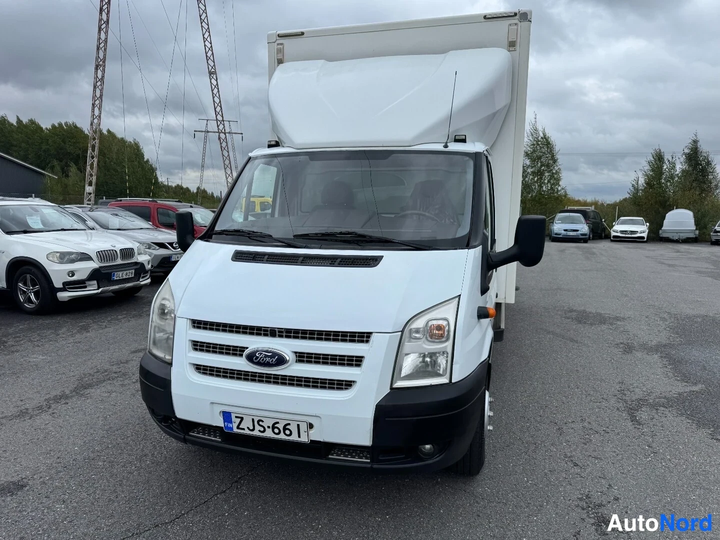 Etuviisto, nokka vasemmalle: Valkoisen Ford Transit -kuormaauton etunurkanäkymä nokka vasemmalla, valkoinen koriboksi, rekisterikilpi.