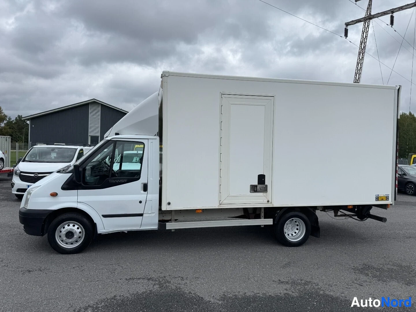Etuviisto, nokka vasemmalle: Valkoinen Ford Transit pakettiauto kylki näkyvissä, keula osittain kuvassa.