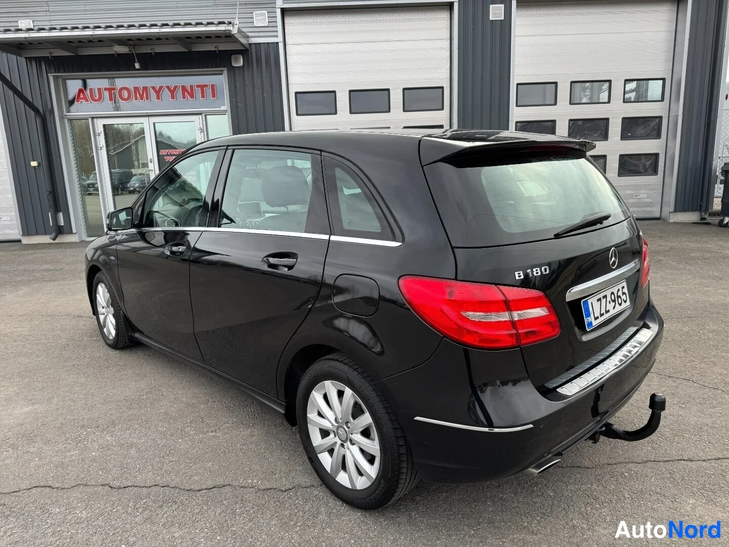 Takaviisto: Mustan Mercedes-Benz B-sarjan takakulmakuva, takana -kilpi ja vetokoukku autoliikkeen edessä.