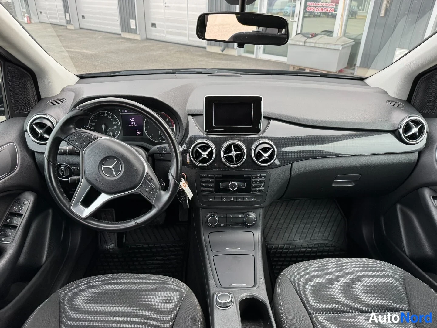 Muu: Mustan Mercedes-Benz B-sarjan ohjaamo: Mercedes-ratti, keskellä kolme pyöreää ilmastointisuutinta, näyttö ja manuaalivaihto-ohjaus, tummat istuimet.