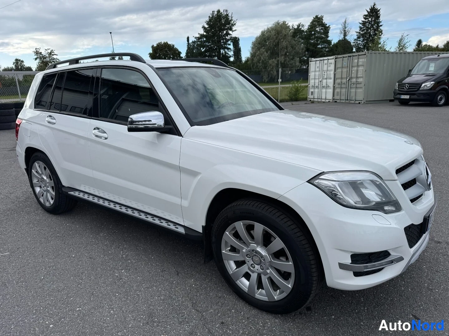 Etuviisto, nokka oikealle: Mercedes-Benz GLK:n etukulmakuva oikealta, valkoinen koriväri, kattokaiteet ja kylkivetoketju näkyvät.