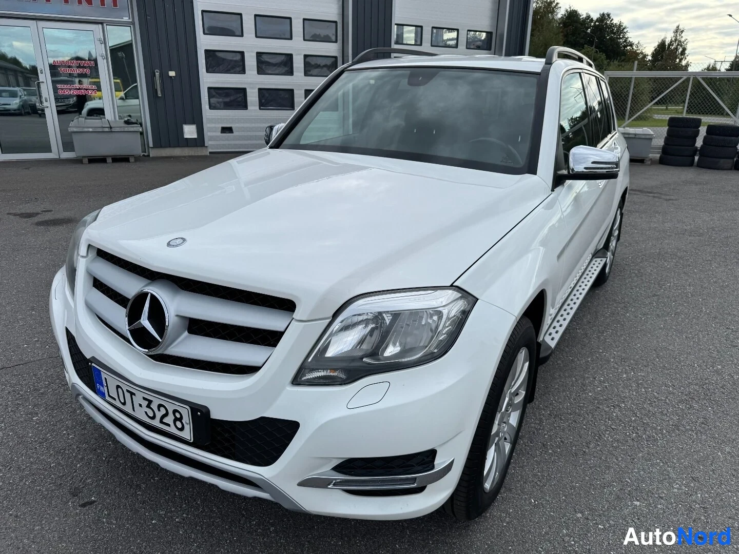 Etuviisto, nokka vasemmalle: Valkoinen Mercedes-Benz GLK -maasturia etuviistöstä vasemmalta, etupanssarissa rekisterikilpi, kattołatvoja ja kirkaskromattuja peilejä.