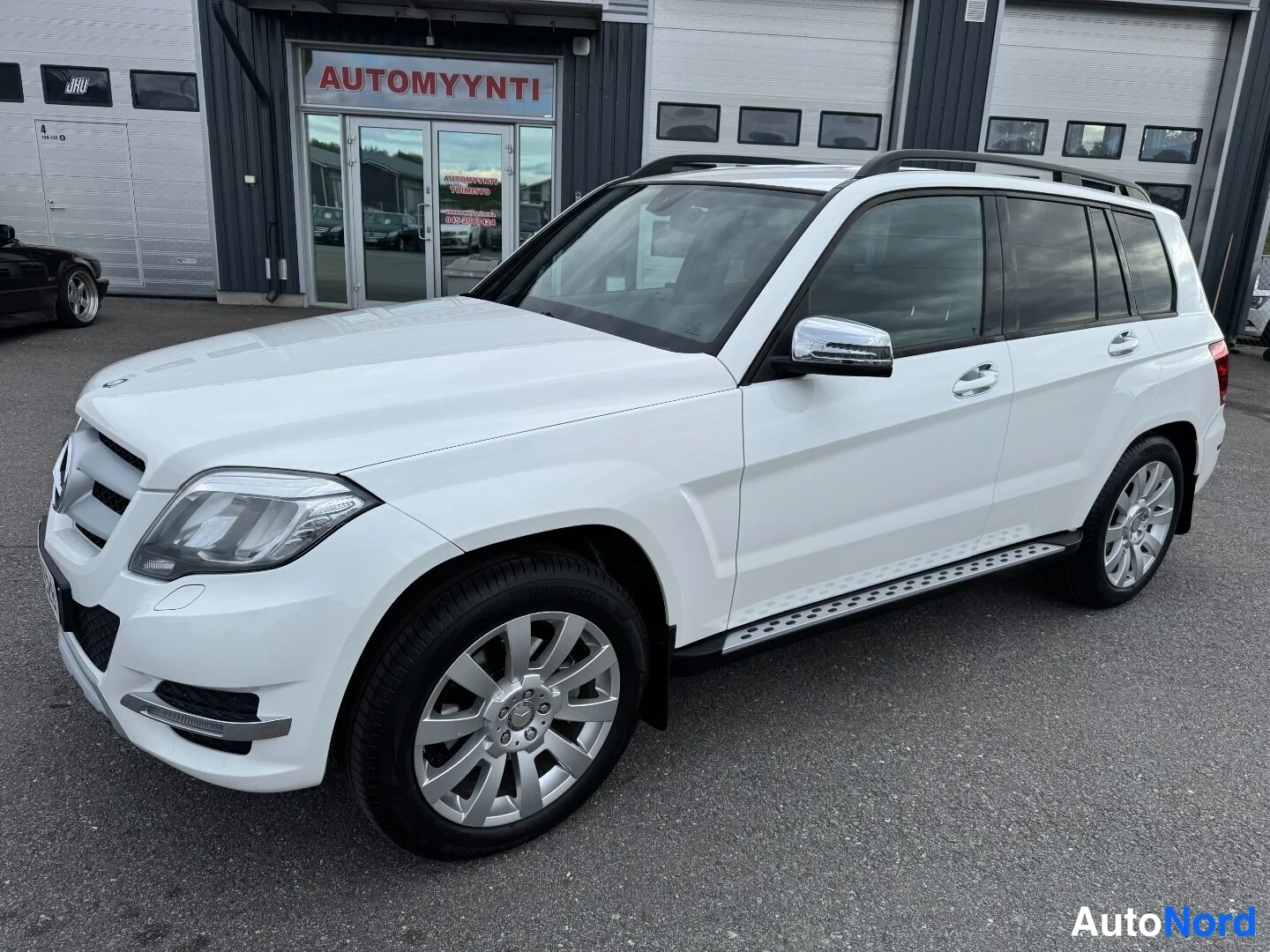 Etuviisto, nokka vasemmalle: Valkoinen Mercedes-Benz GLK -maasturi etuviistöstä vasemmalta automyynnin tilojen edessä, kokonaisuus hyvin näkyvissä.