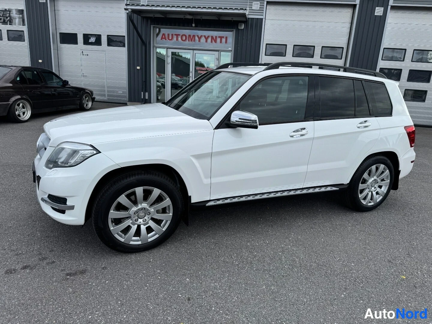 Etuviisto, nokka vasemmalle: Valkoinen Mercedes-Benz GLK -maasturi etuviistöstä vasemmalta, kylki ja keula näkyvissä, kattotelineet asennettu.