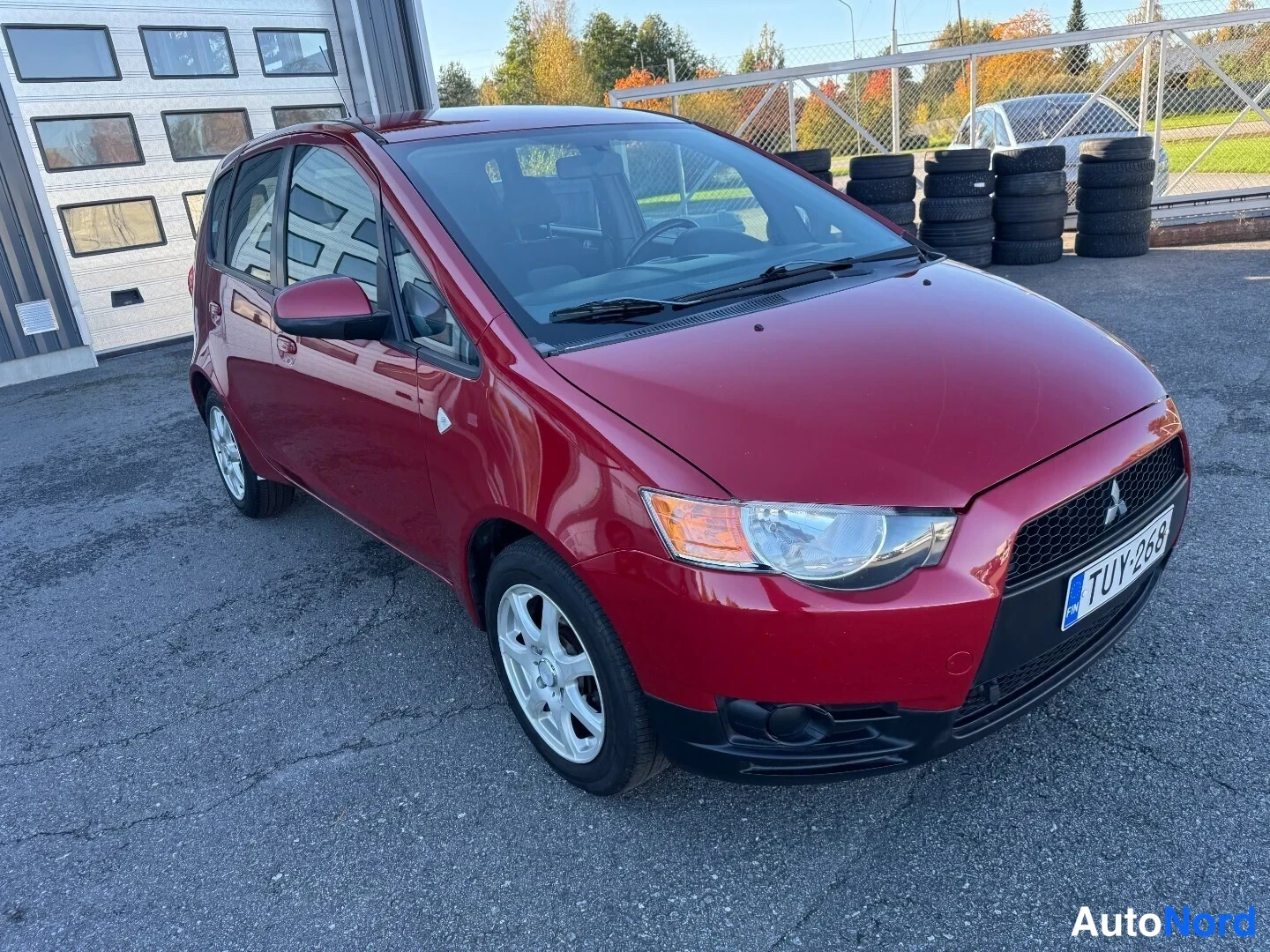 Etuviisto, nokka oikealle: Punainen Mitsubishi Colt etuviistosta oikealta, rekisteritunnus, auton vieressä pinoja rengaita.