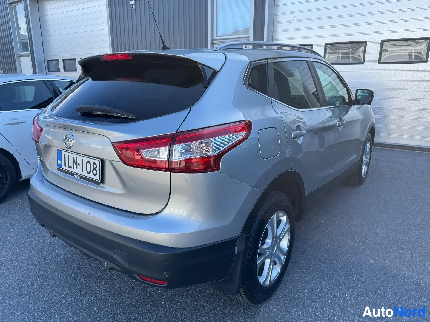 Takaviisto: Hopeanharmaa Nissan Qashqai takaa ja oikealta sivulta kuvattuna, takaosan pyyhkija ja rekisteritunnus näkyvät.
