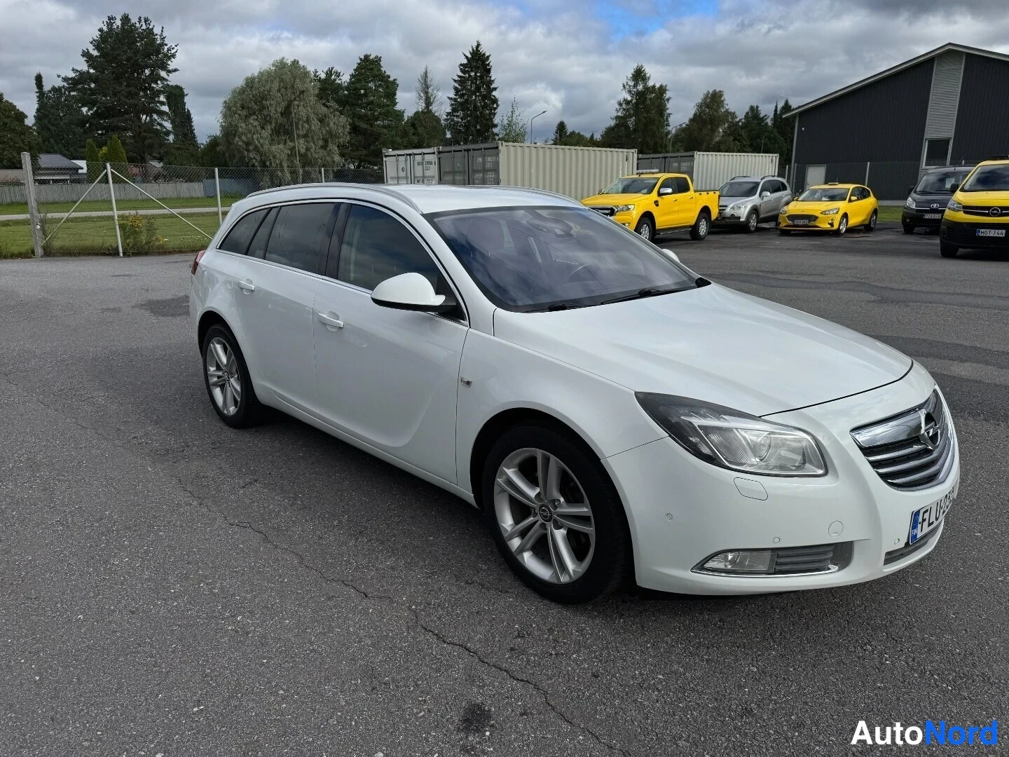 Etuviisto, nokka oikealle: Valkoisen farmariauto etuapäältä oikealta kuvattu; etupyörässä kevytmetallivante, konepellissä Opel-merkki ja etulokasuojassa -rekisterikilpi.