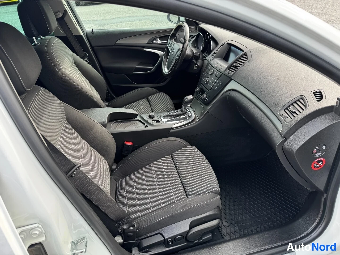 Muu: Opel Insignian etusohva ja kojelauta kuvattuna avoimesta etiovesta; tummat kankaanistuimet, ruoripöytä ja keskikonsoli näkyvät.