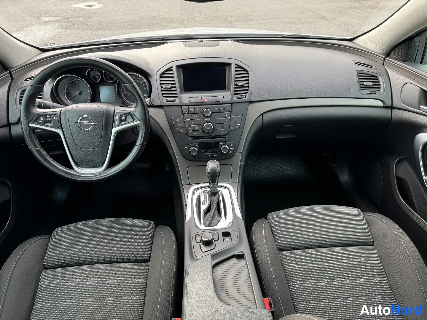 Muu: Ohjaamo edestä: Opel-merkkinen ohjauspyörä, tumma kojelauta, keskikonsolissa infotainment-näyttö ja automaattivaihteiston valitsin, kaksi tummaa kangasistuinta.