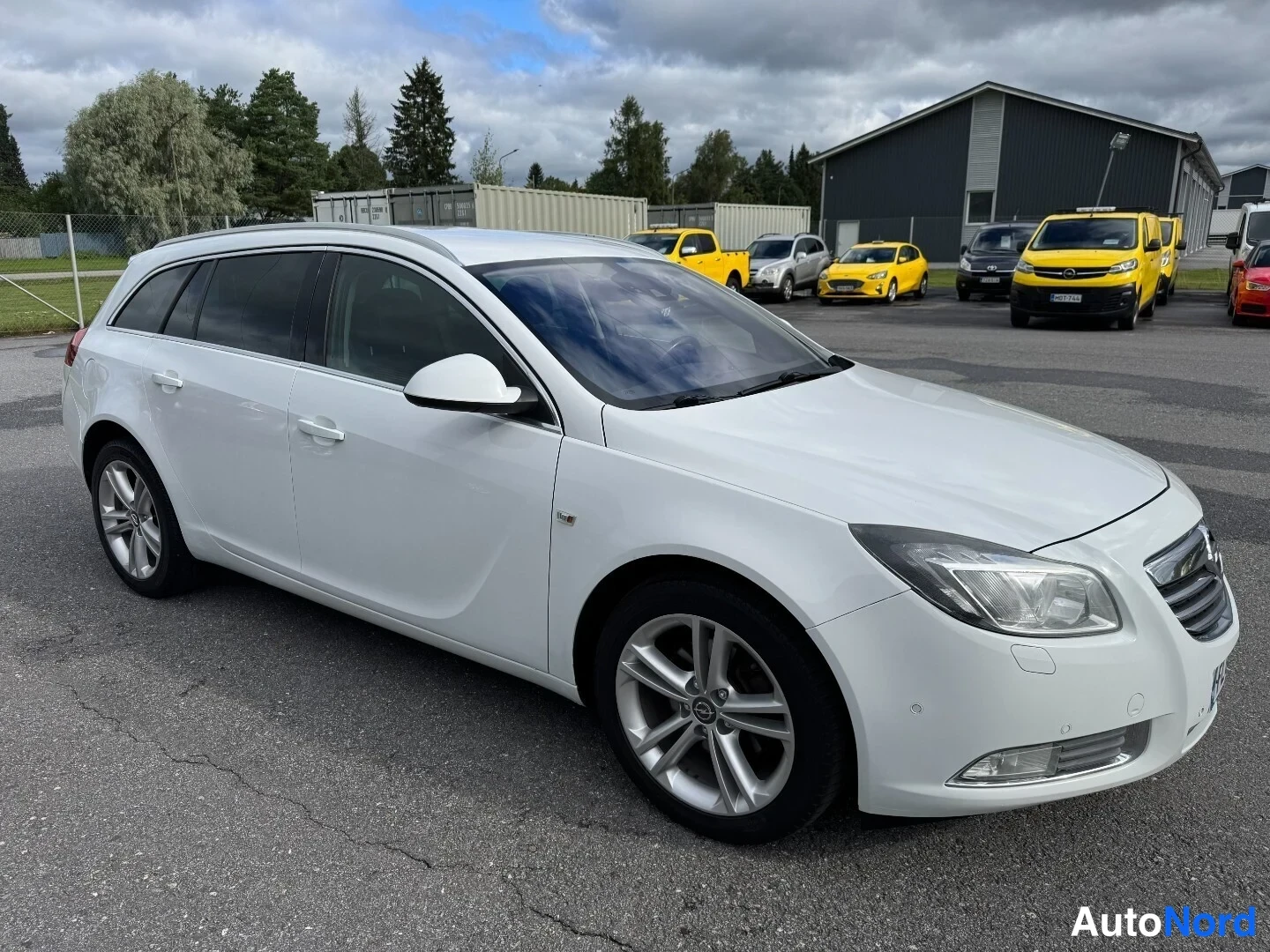 Etuviisto, nokka oikealle: Valkoinen Opel Insignia -farmari kolmasosaedestä oikealta, etusäleikkö ja ajovalot sekä oikea kylki näkyvissä parkkipaikalla.