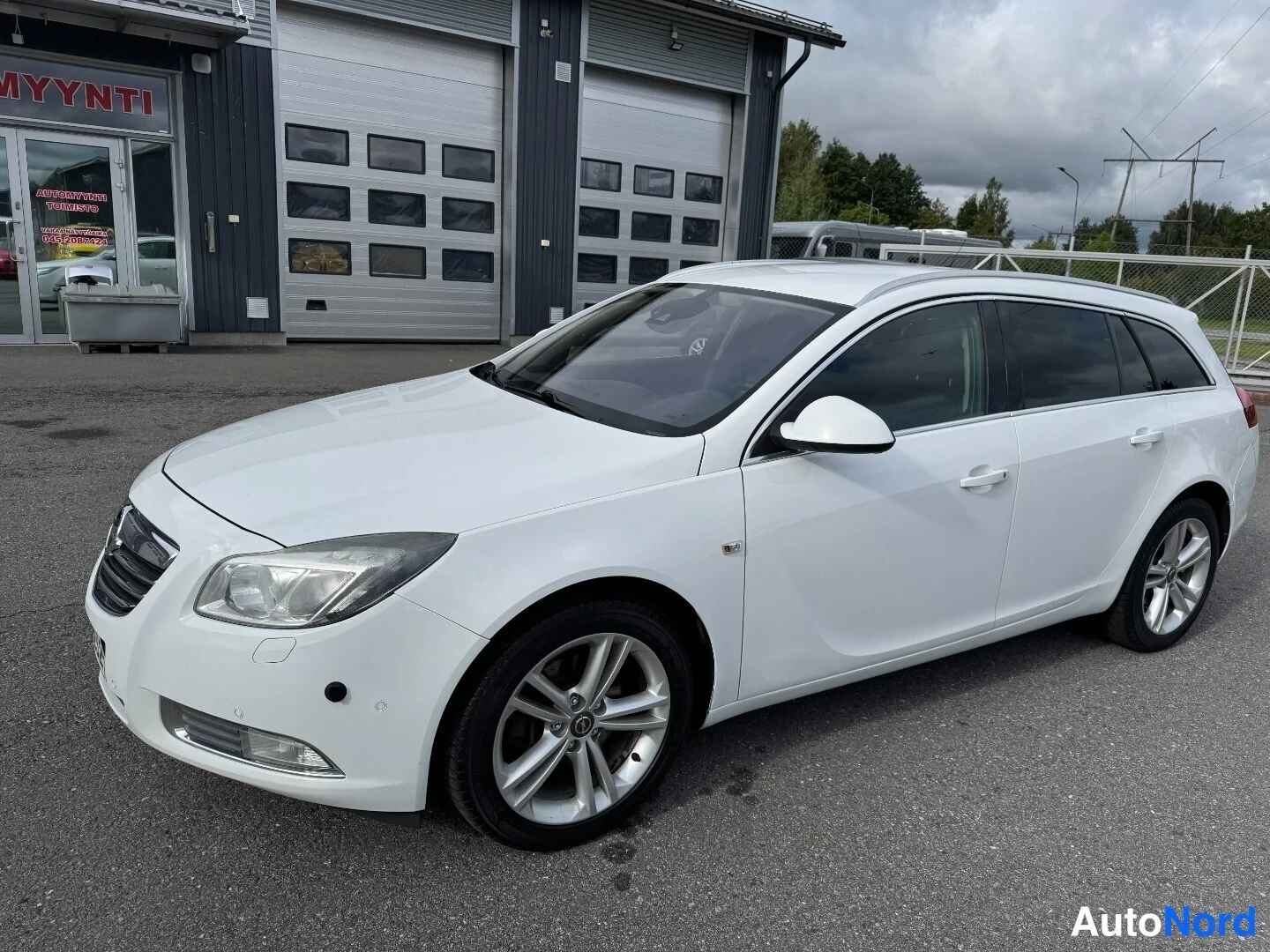 Etuviisto, nokka vasemmalle: Valkoisen Opel Insignia -farmarin vasen sivukulma; kylki, peilit ja hopeat alumiinivanteet näkyvät autopihan edessä.