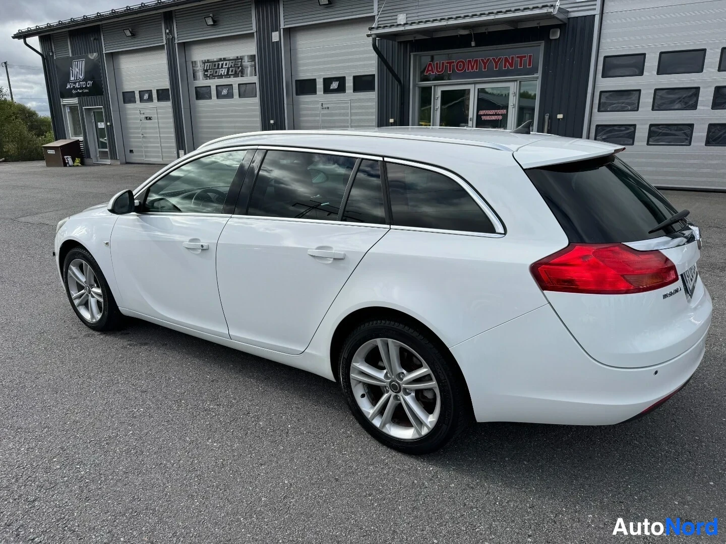 Takaviisto: Valkoisen Opel Insignia -farmarin takakulma; punaiset takavalot, takaluukku ja takaveto näkyvät korjaamorakennuksen edessä.