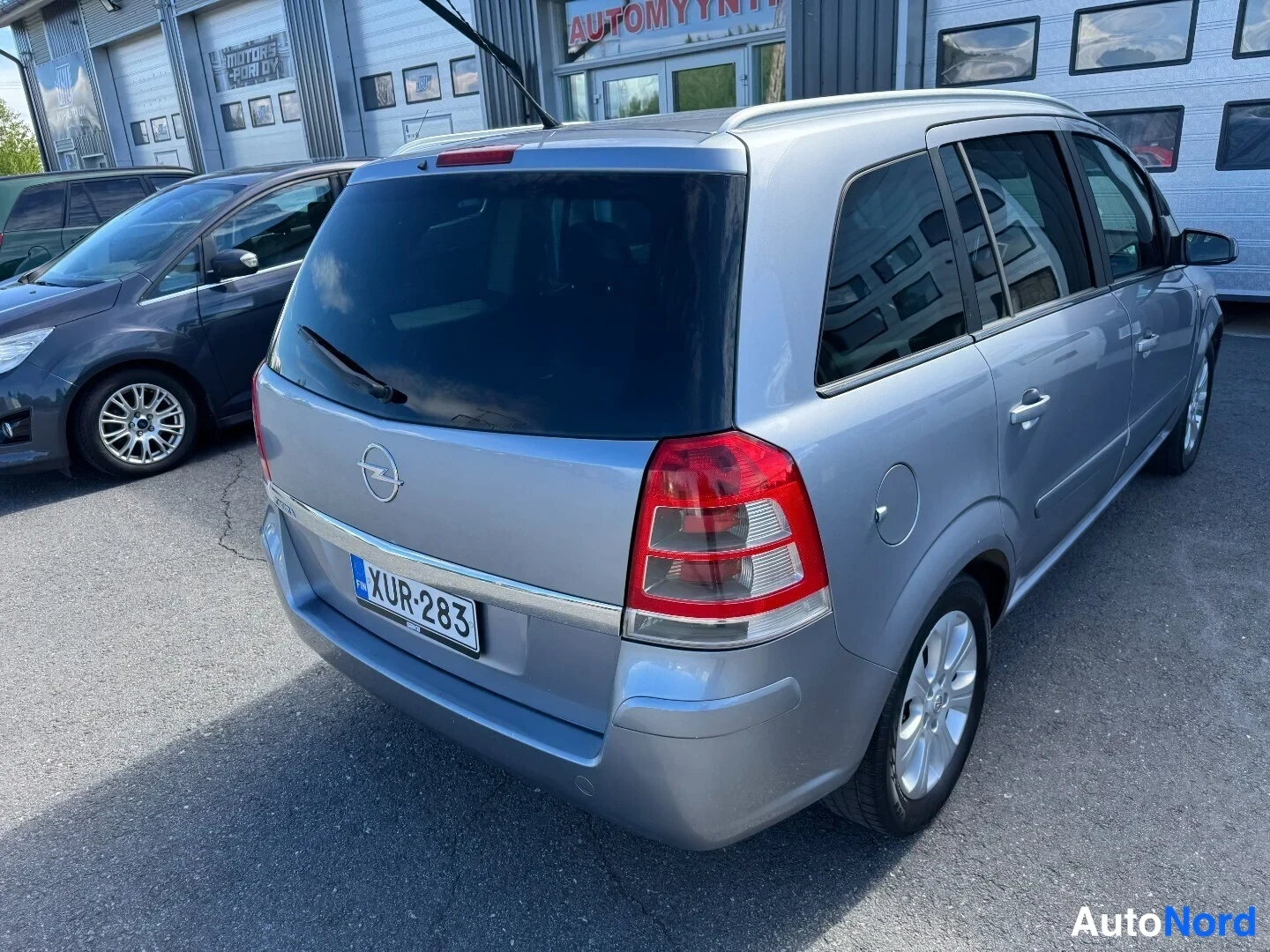 Takaviisto: Hopeanharmaa tila-auto vinosti takaa sivulta kuvattuna, takamoottorin tassut ja oikea kylki näkyvät, rekisteritunnus.
