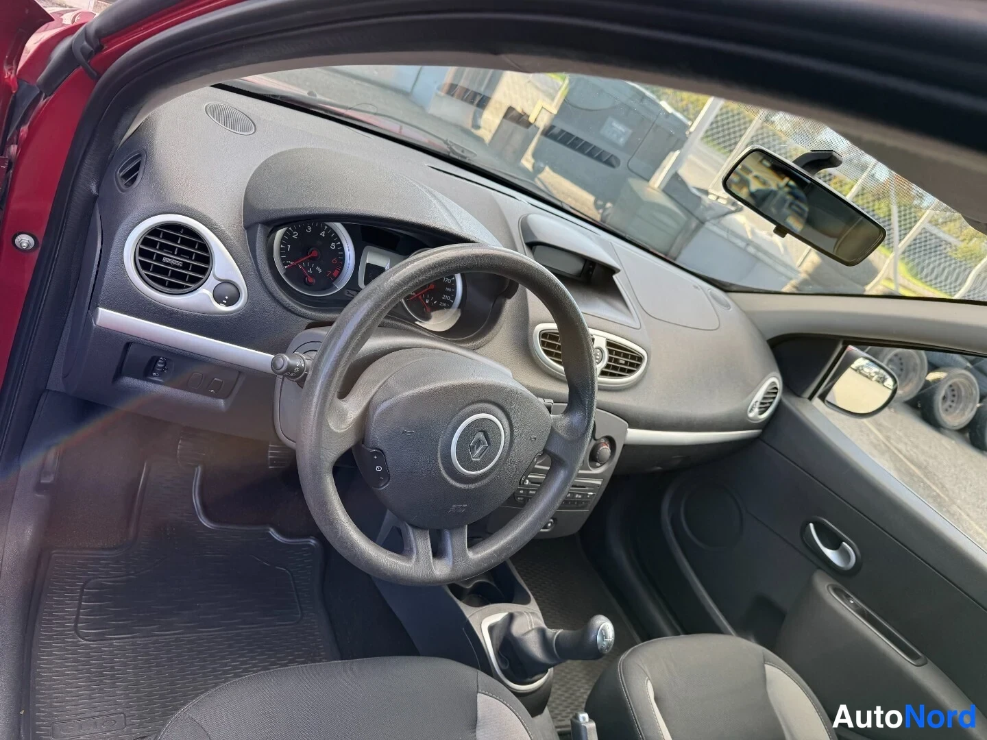 Muu: Renault Clio -matkailuauton sisätilat: tumma kojelauta, Renault-ohjauspyörä, analogiset mittarit, manuaalivaihteen keksi ja kaksi etutiltaa kangasverhoiluilla.
