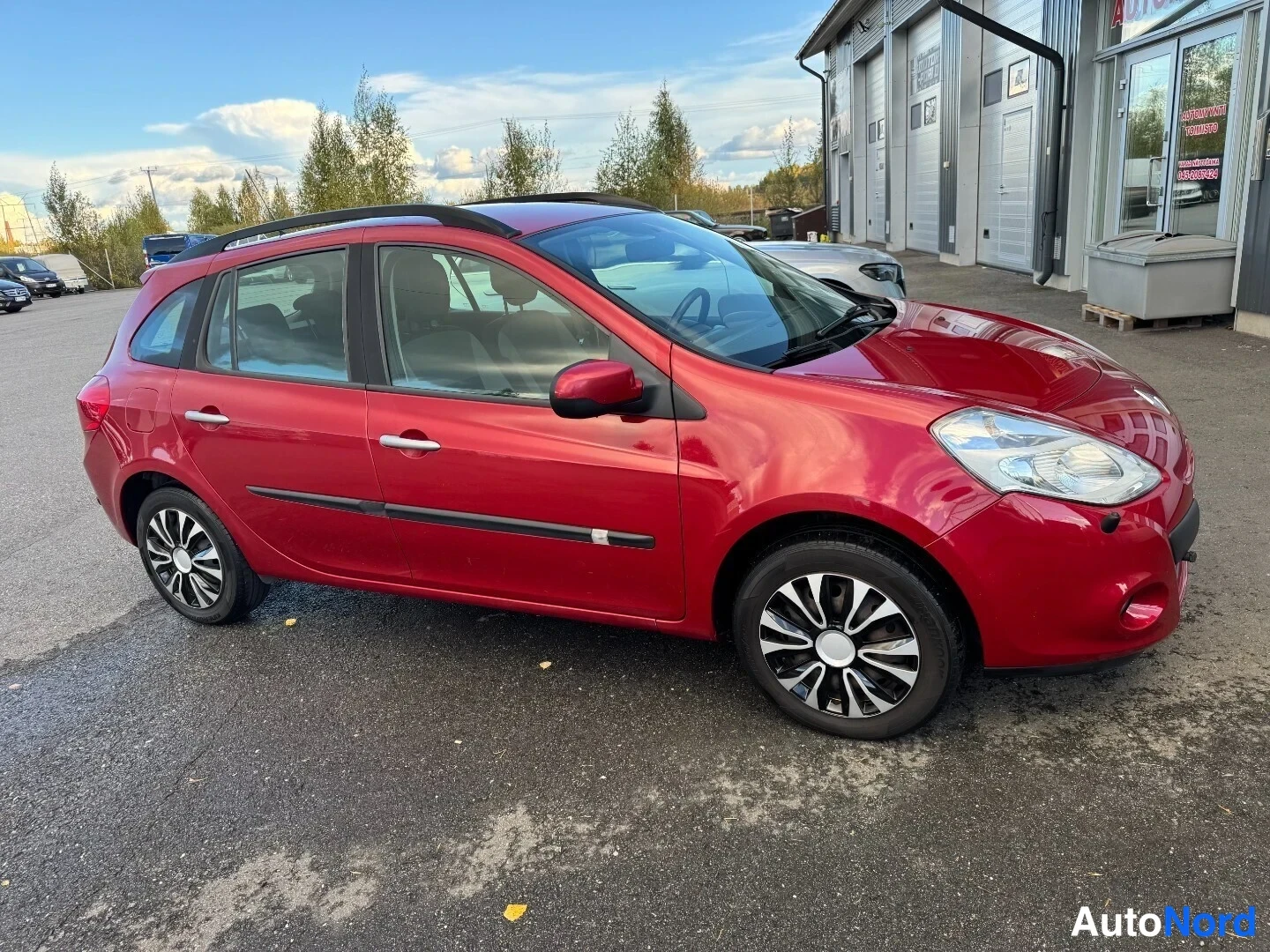 Sivulta: Punainen Renault Clio farmari sivulta vasemmalta, kattokaiteet ja moniosaiset kevytmetallivanteet näkyvät.