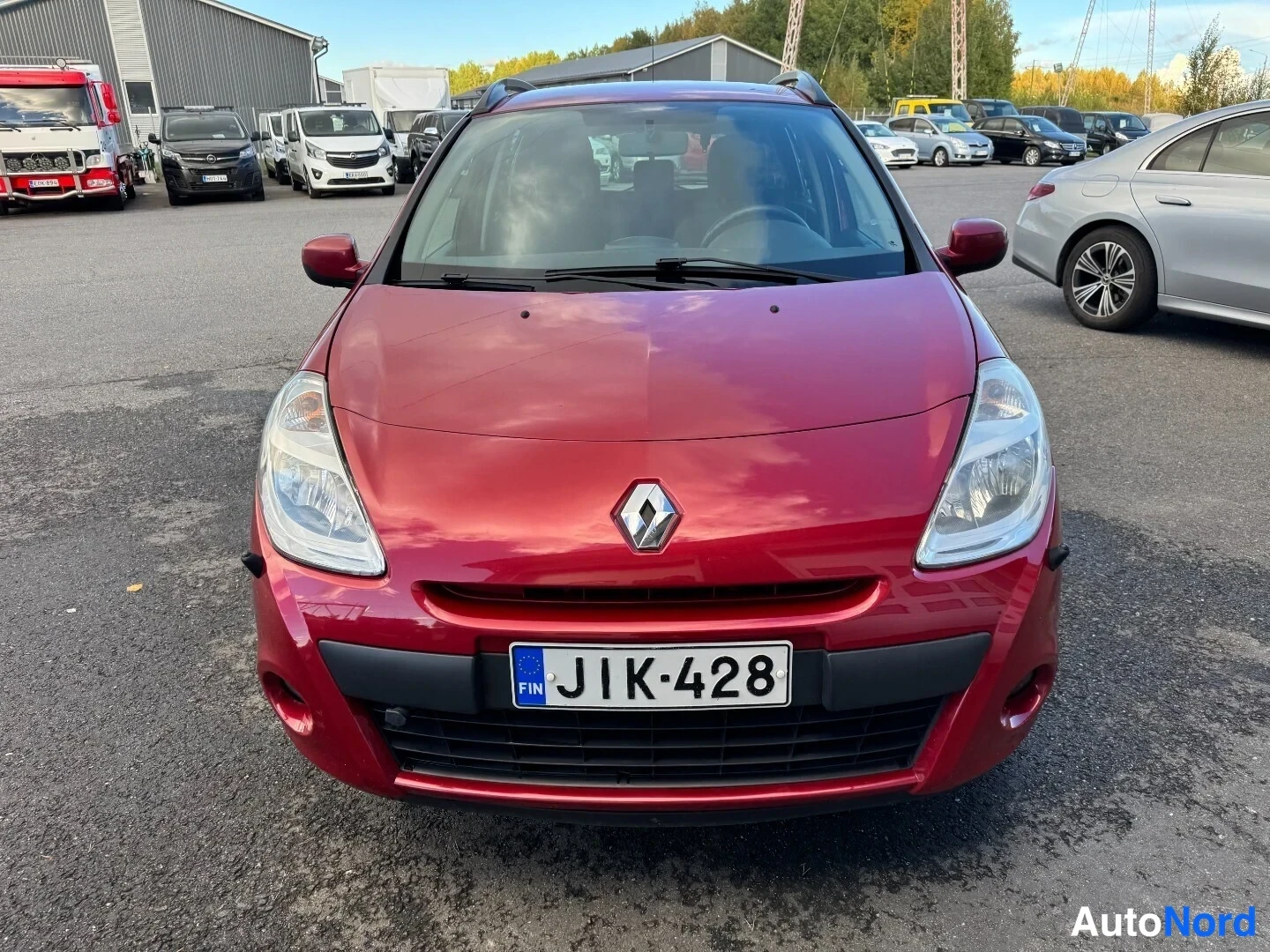 Edestä: Punaisen Renault Clio Tourerin etupuoli suoraan edestä valokuvaamossa; kylkikiila näkyy Rekisterikilpi on kiinnitetty etupuskuriin. Etuvalot, Renault-merkki ja